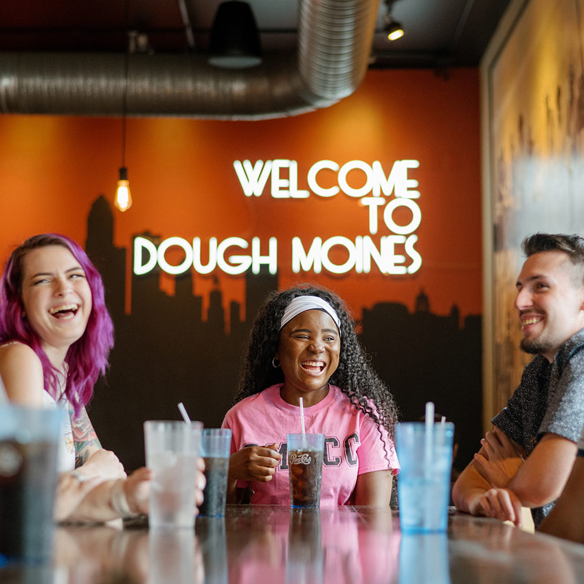 people sitting and talking with a sign in the background that states Welcome to Dough Moines